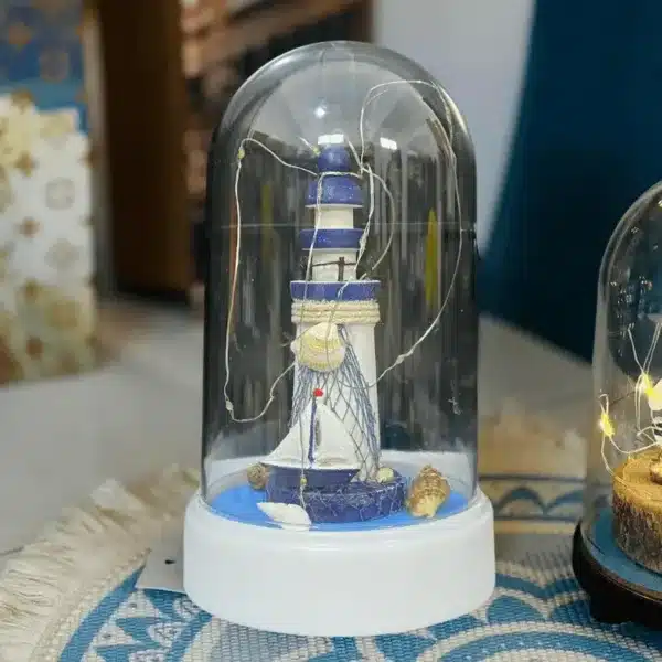 Lampe décorative marine en forme de phare sous cloche en verre avec une mouette et des coquillages, accompagnée d’une boîte à thème océan.