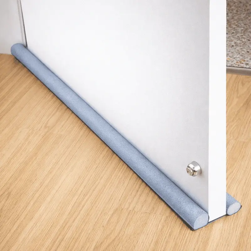 bas-de-porte-etanche-anti-froid-et-poussiere-gris Bas de porte étanche anti froid glissé sous une porte blanche sur sol en bois