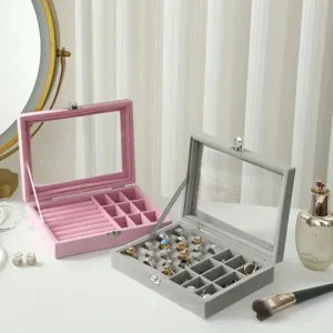 Boîte à bijoux en velours rose et gris avec couvercle en verre sur une coiffeuse.