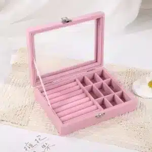 Boîte à bijoux en velours rose et gris avec couvercle en verre sur une coiffeuse.